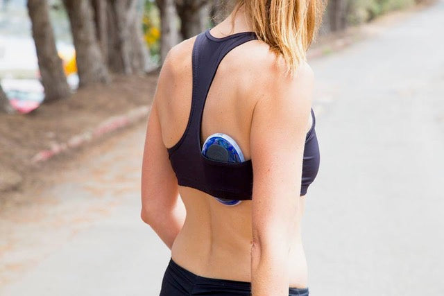 A woman from behind wearing a black sports bra with a blue and white pain management device on her back, standing on an outdoor paved path.